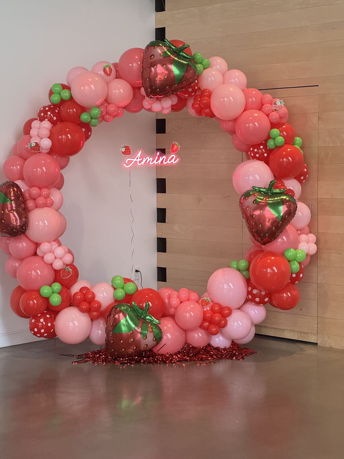 A red, pink, and green strawberry-themed round balloon arch with a neon Amina sign